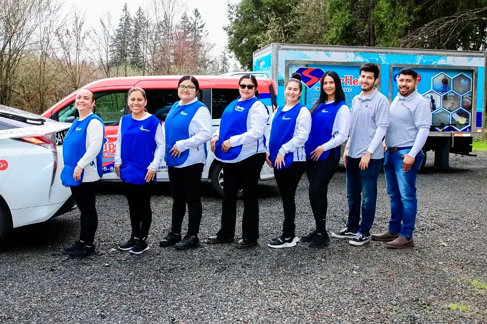 Eight Superhero Cleaning team members, most in blue aprons, lined up in front of the branded company van and car