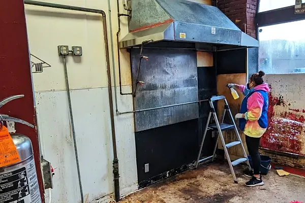 A crew member on a step stool spraying and wiping a grease-stained commercial kitchen wall under a range hood