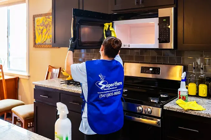 Superhero Cleaning team member in the blue company apron wiping down a microwave above the stove